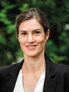 Elena Hassinger draußen, mit Blazer und Ohrringen, lächelnd.